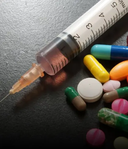 A syringe filled with liquid and varies pills and tablets scattered around a table.