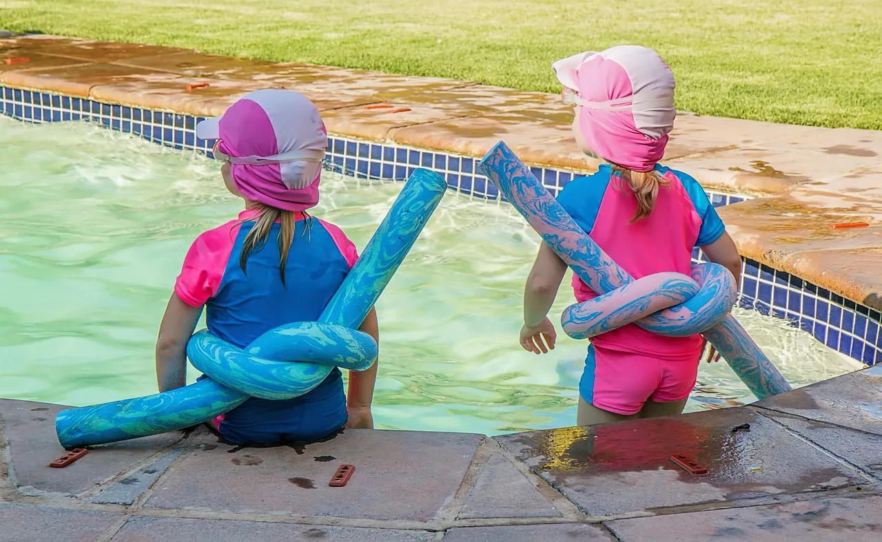 Two children sit by a pool wearing flotation devices.
